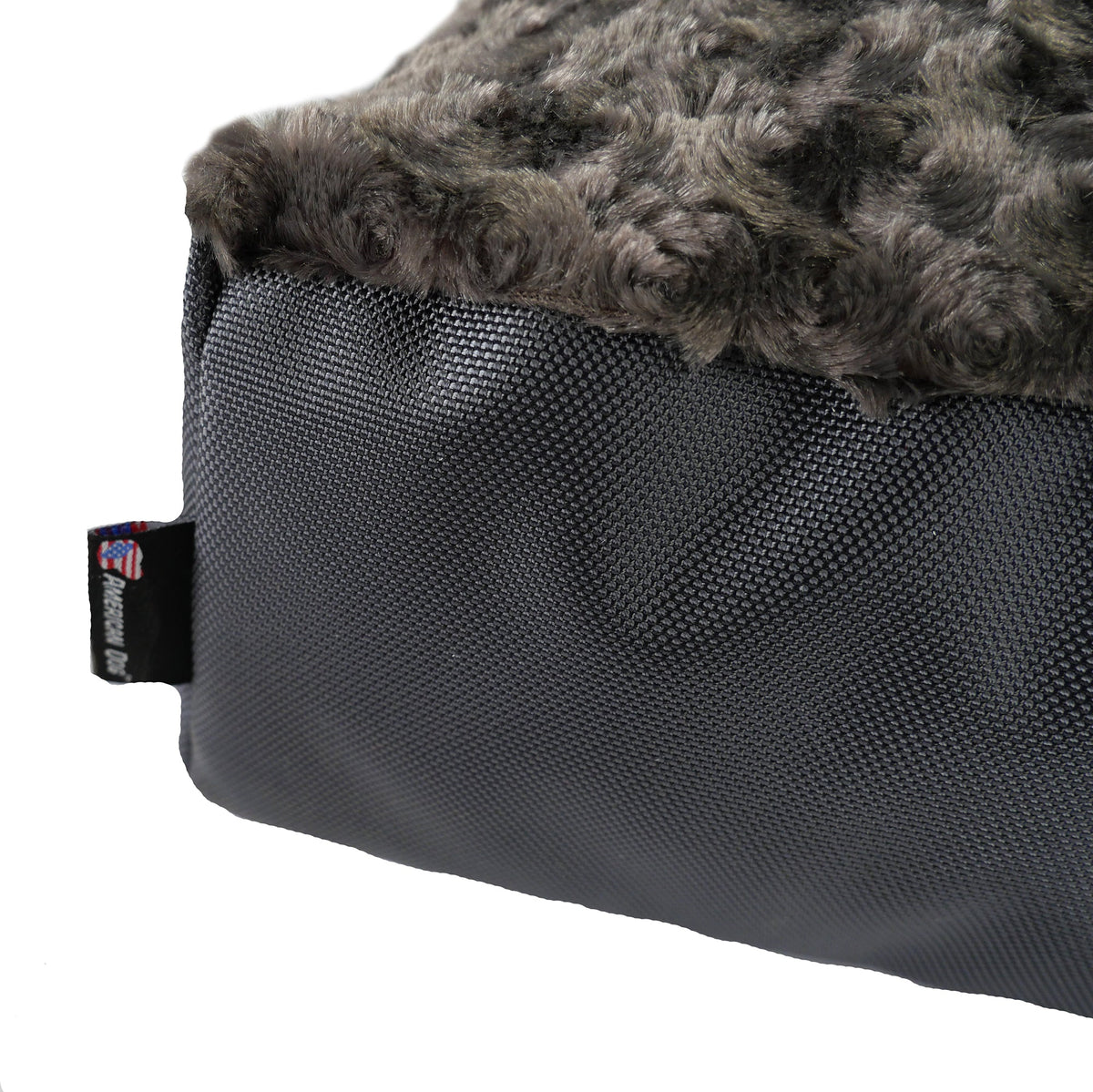 Corner close-up of a dog bed showing charcoal plush top over gray ballistic nylon side and brand tag