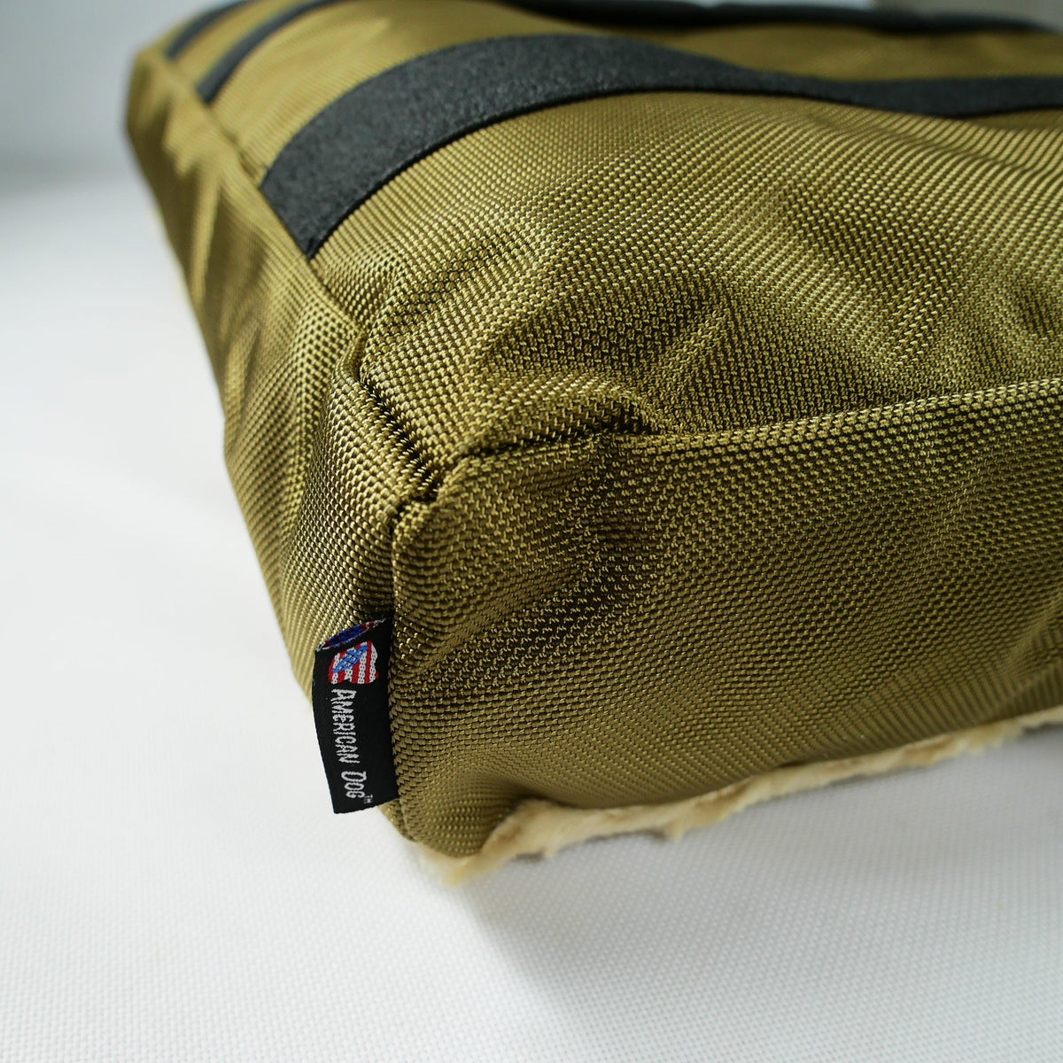 Corner close-up of a dog bed showing olive gold ballistic nylon side, black strap and woven brand tag